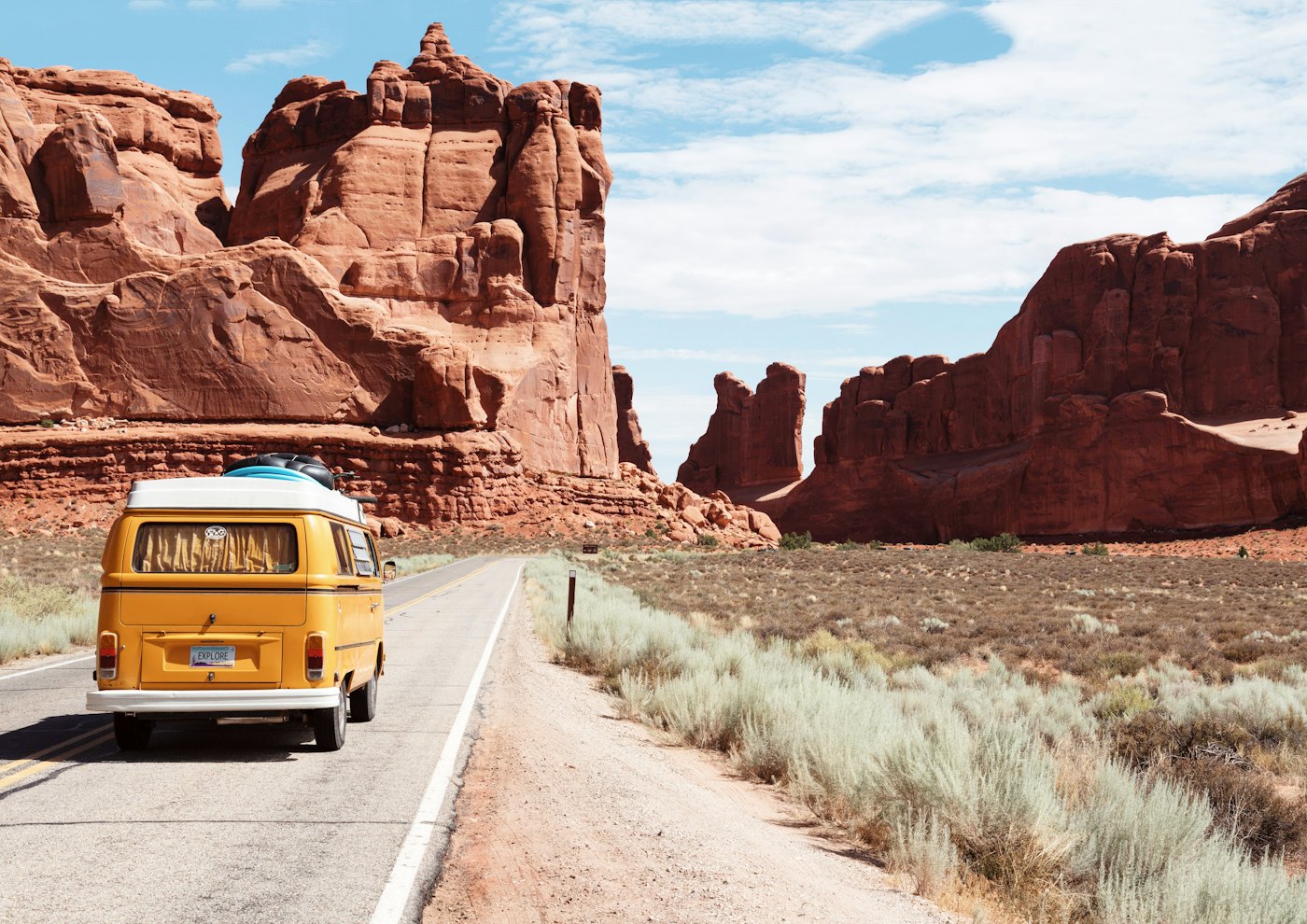 A yellow van driving away on a desert highway between red rock walls.