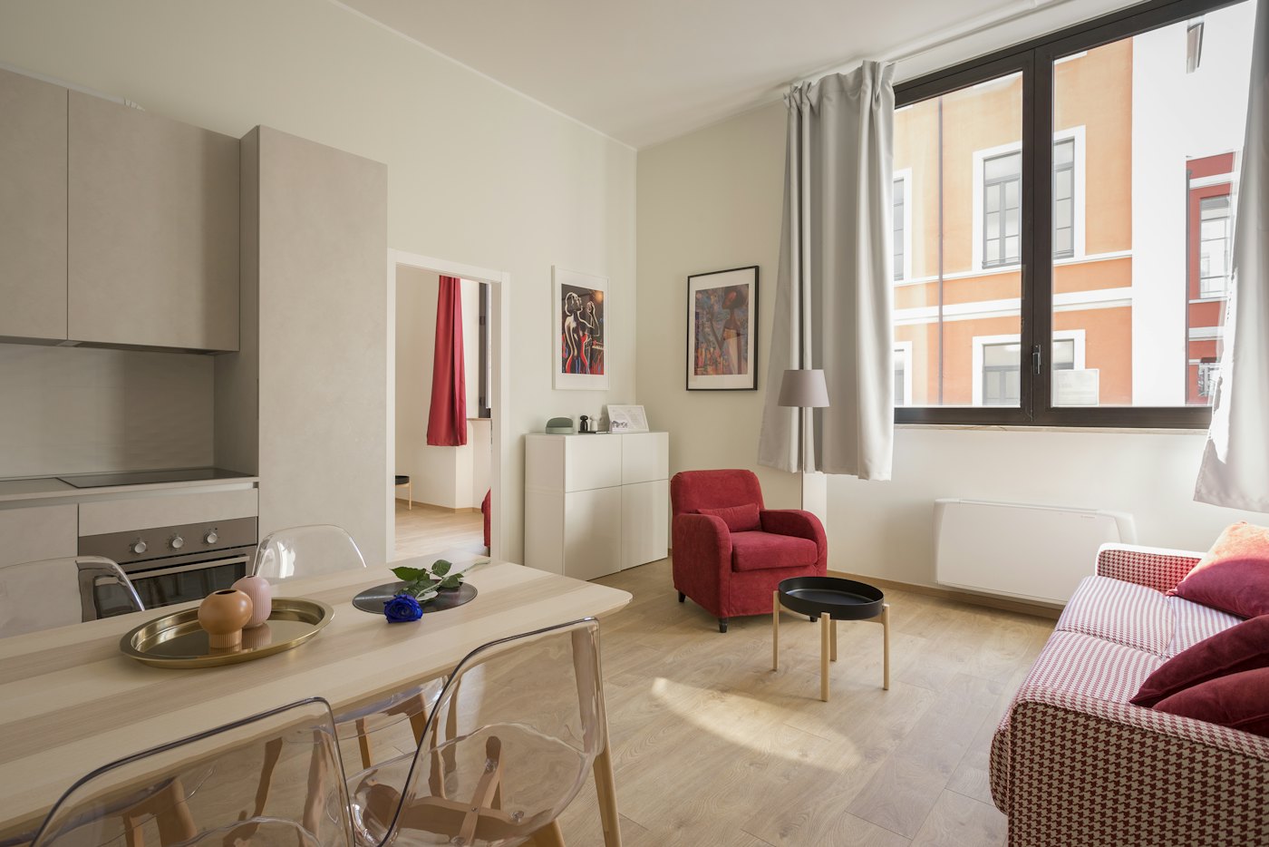 A sunlit apartment room with a red chair, a pale sofa, and a large window.