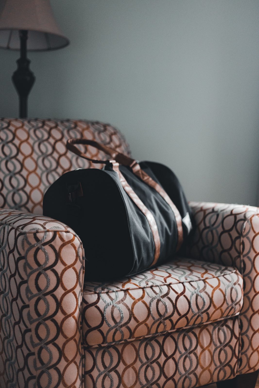 A closed black duffel with brown straps resting on a patterned armchair, with a lamp against a plain wall behind it.
