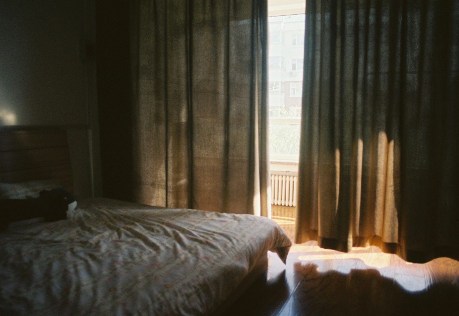 A rumpled bed beside heavy curtains that do not quite meet, with warm light falling across the wooden floor and a radiator visible under the window.